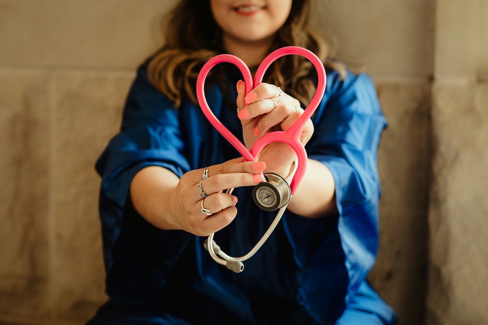 A smiling healtchare worker in blue scrubs holds out a bright pink stethoscope shaped like a heart.